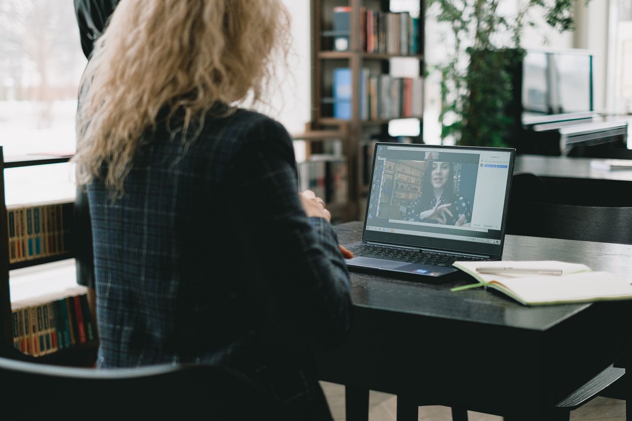 A woman is seated at a laptop in an elegant library environment, taking part in a video call.